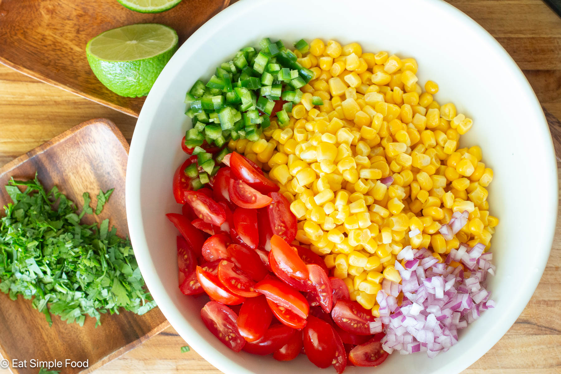 Top down view of unmixed ingredient in a white bowl: corn, quartered cherry tomatoes, diced red onions, diced jalapenos. Half of a lime and chopped cilantro on the side.