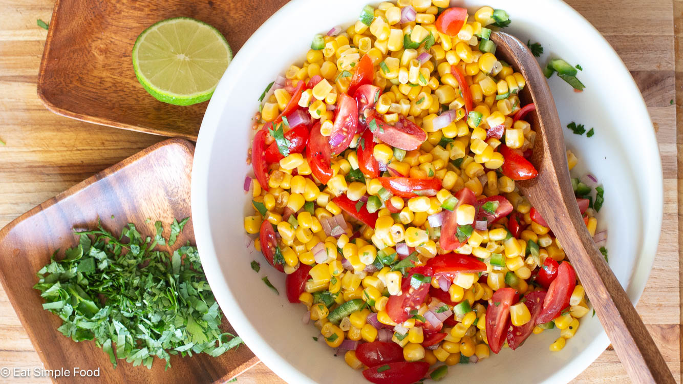 Top down view of mixed ingredient in a white bowl: corn, quartered cherry tomatoes, diced red onions, diced jalapenos. Half of a lime and chopped cilantro on the side.