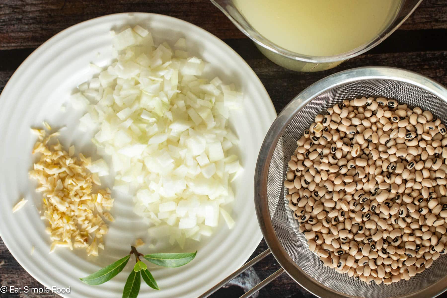 Top down view of white beans with a black spot in a silver colander. White plate on the side with diced onions, minced garlic, and bay leaves.