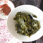 Top down view of white bowl with chopped greens and sauce in it. Small bowl of chili flakes on the side.