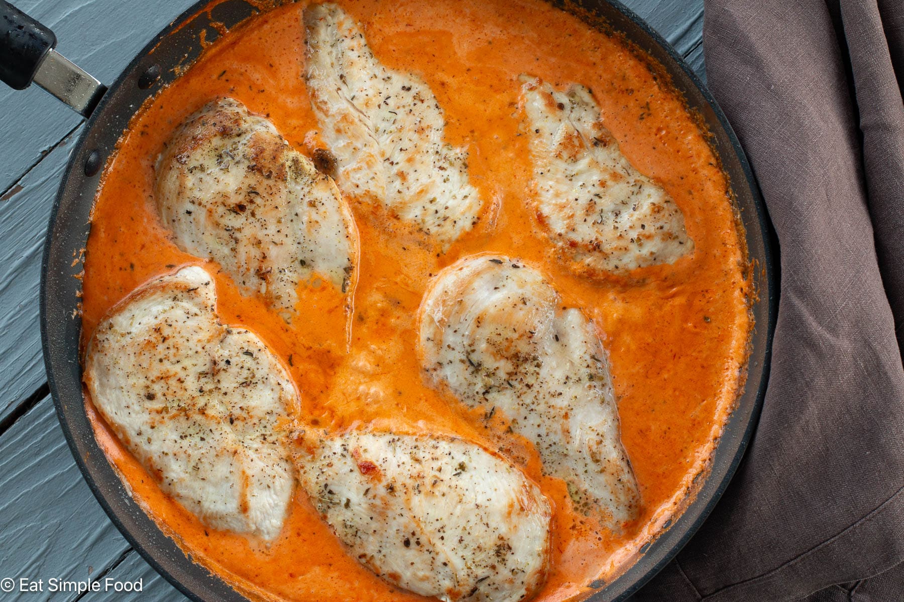 Top down view of six pieces of seared chicken breast in a red sauce in a black pan with a brown napkin on the side.