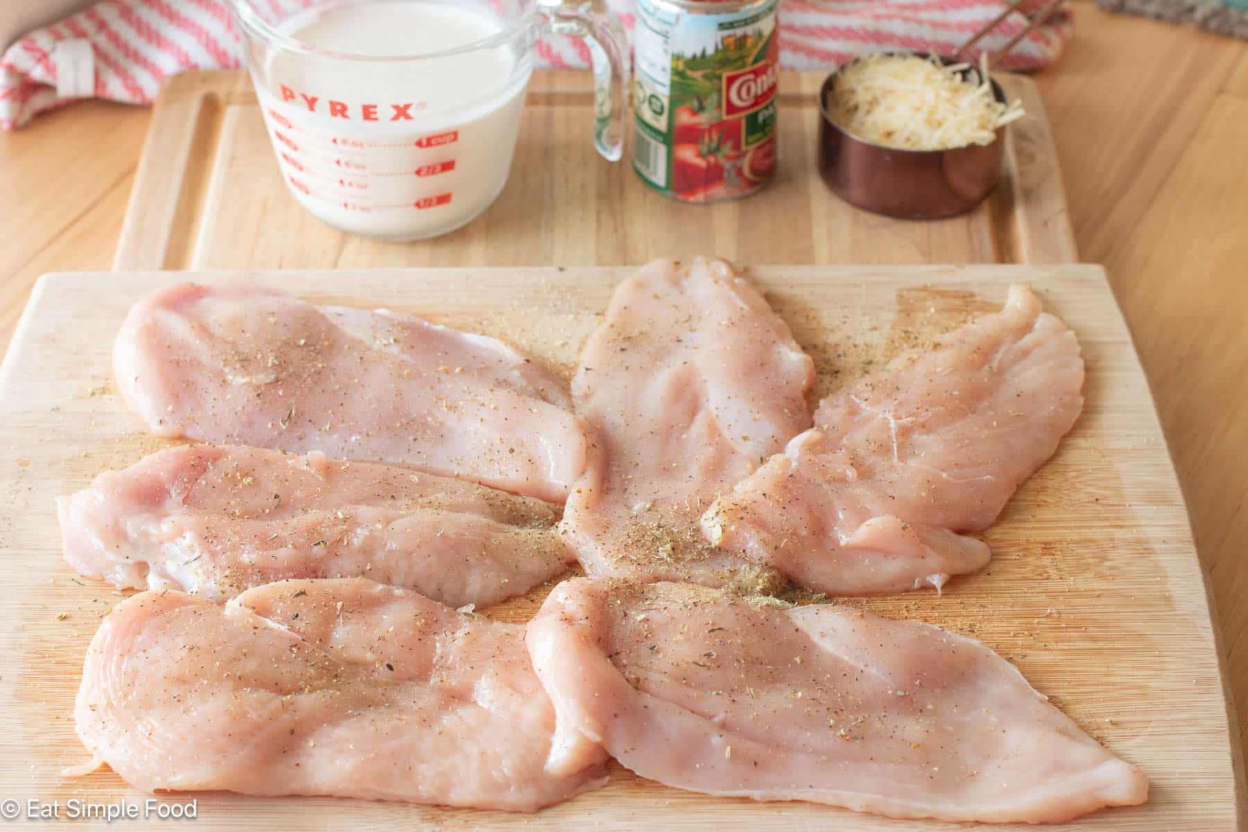 6 pieces of raw chicken on wood cutting board that have spices on them. Ingredients in background.