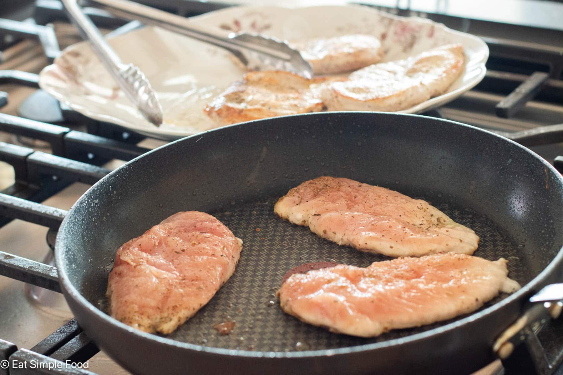 Three thin pieces of raw chicken cooking in a black pan on the stovetop. Plate in background.