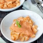 Close up of white plate with chicken and noodles in an orange sauce with basil leaves on top.