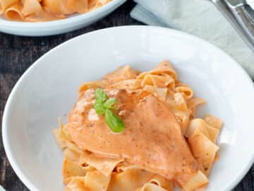 Close up of white plate with chicken and noodles in an orange sauce with basil leaves on top.