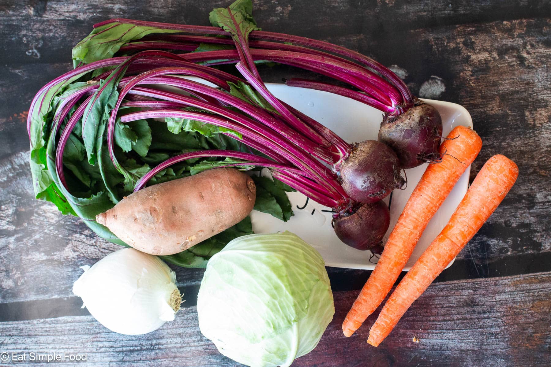 Ingredients on a white plate on a dark wood table? A bunch of beets, a sweet potato, a head of cabbage, one onion, and two carrots.