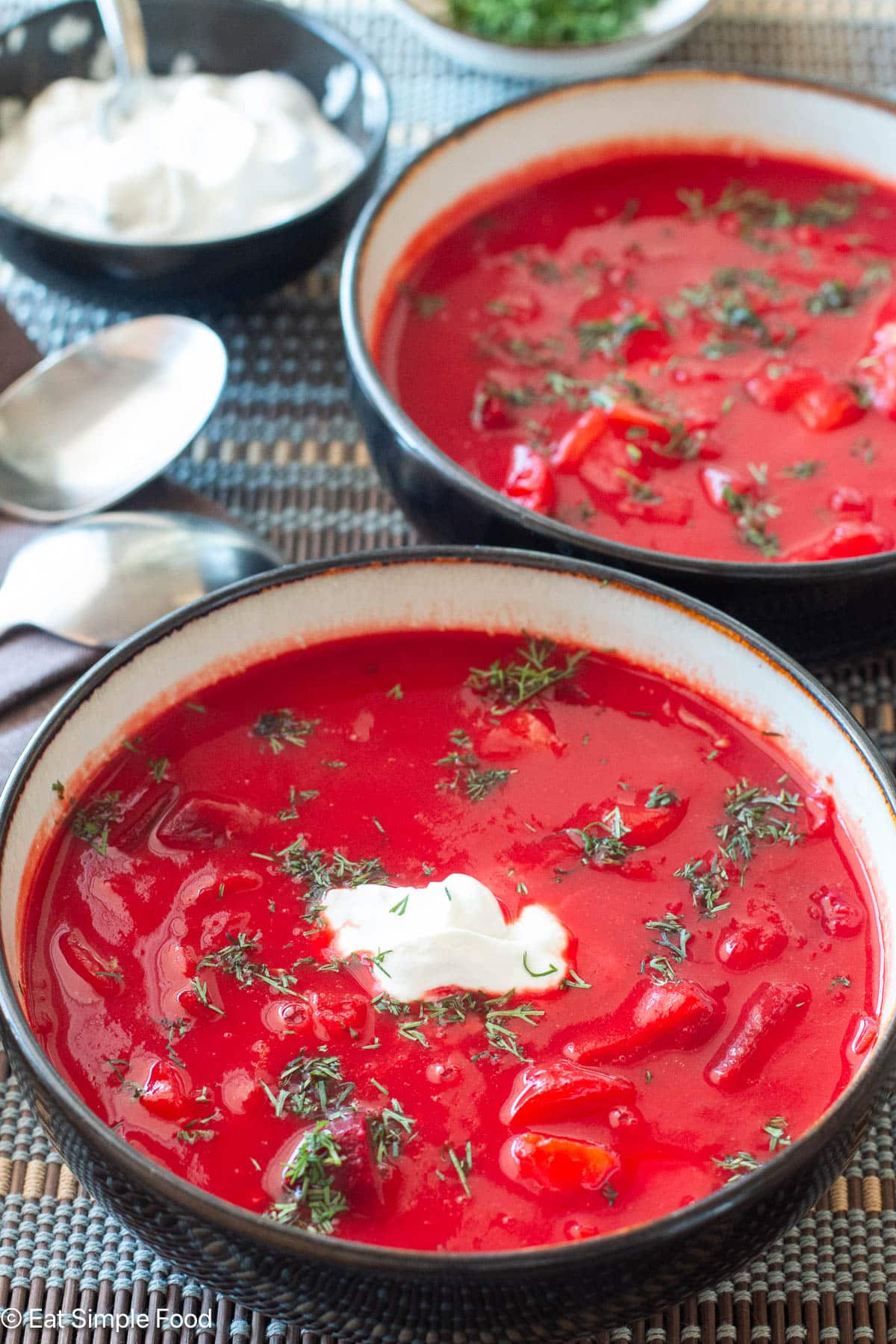 Side view of two bowls of red soup topped with dill and sour cream. Purple napkins and spoons on the side.