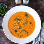White bowl of orange soup with green garnish with blue napkin and silverware on the side. Top View