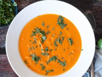White bowl of orange soup with green garnish with blue napkin and silverware on the side. Top View