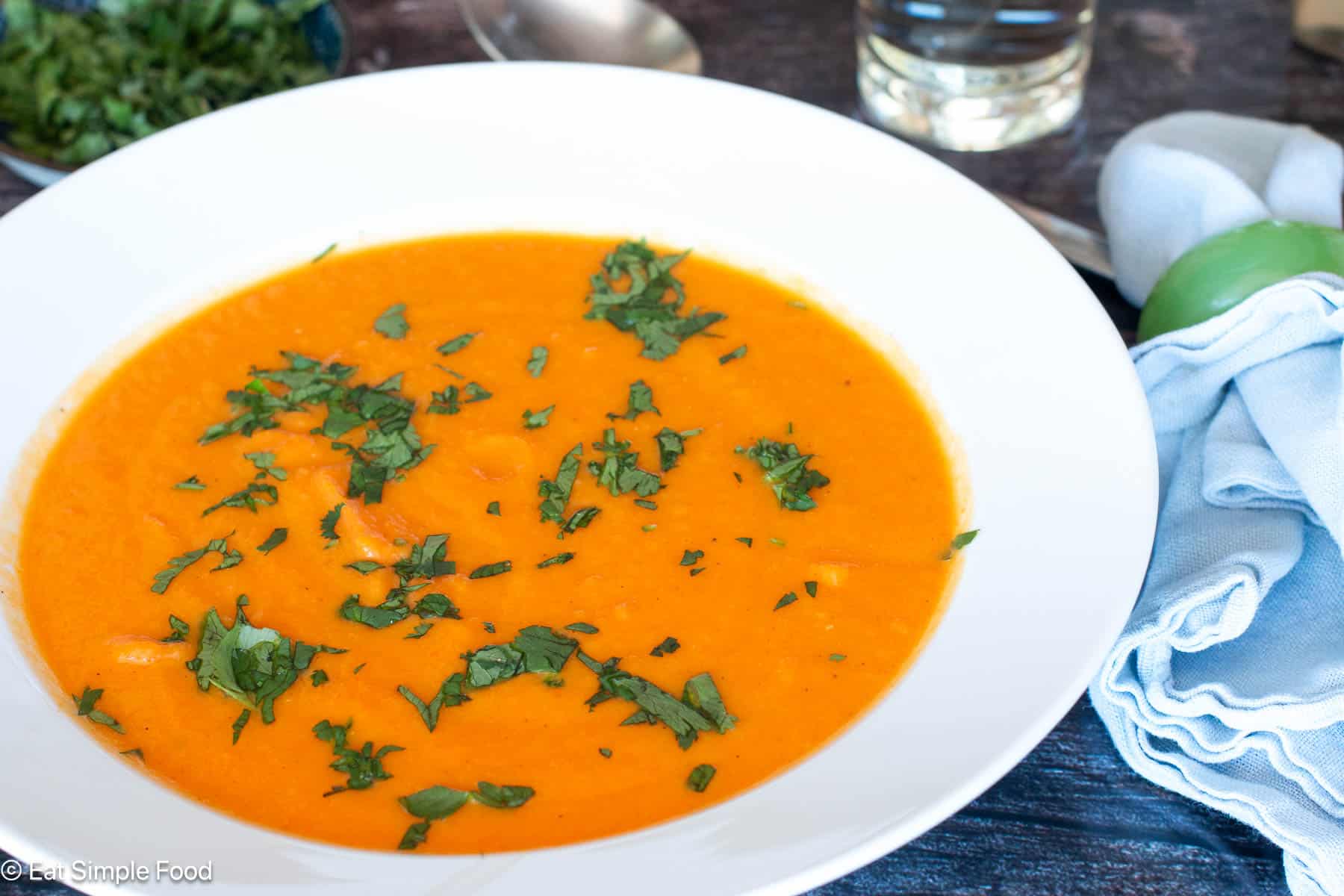 White bowl of orange soup with green garnish with blue napkin and silverware on the side. Side View