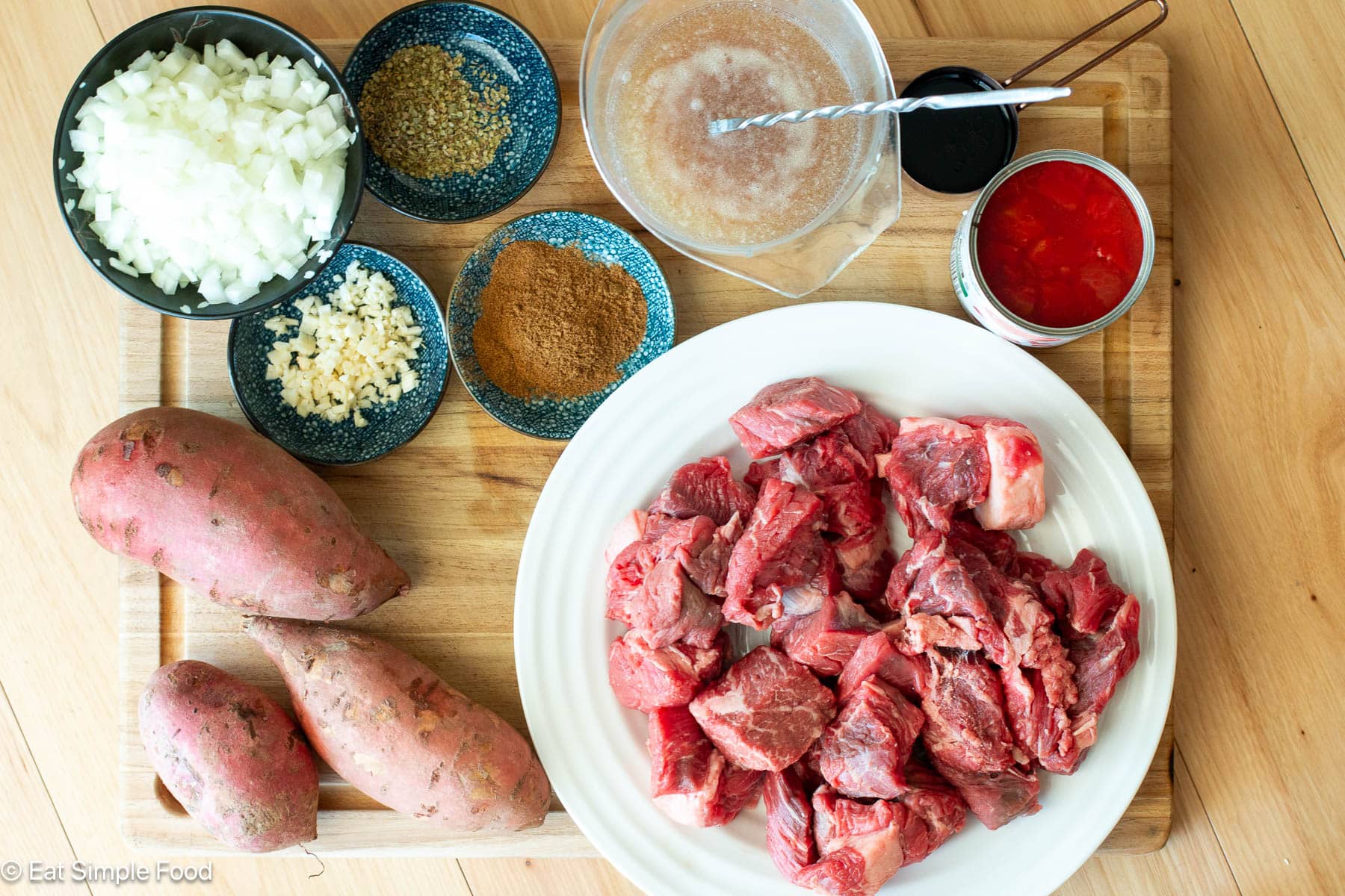 Wood cutting board with ingredients: cubed chuck roast on white plate, canned diced tomatoes, bowl of diced onions, 3 sweet potatoes, and spices.