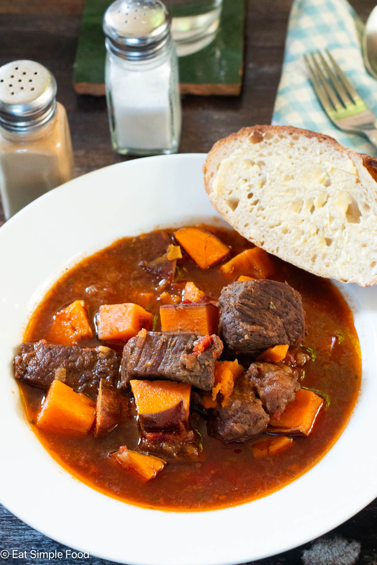 Side view of white bowl of brown stew with chunks of orange sweet potatoes. Piece of buttered bread on side. Salt and pepper shaker in the back.