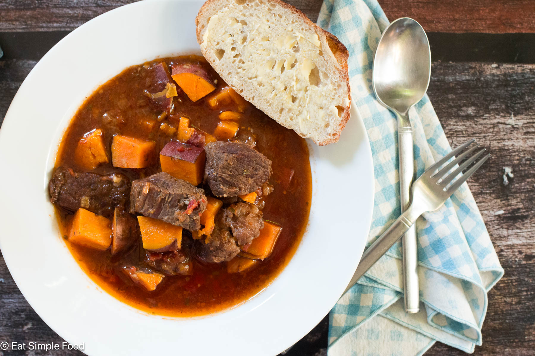 Top down view of white bowl of brown stew with chunks of orange sweet potatoes. Piece of buttered bread on side. Napkin, fork and spoon on the side.
