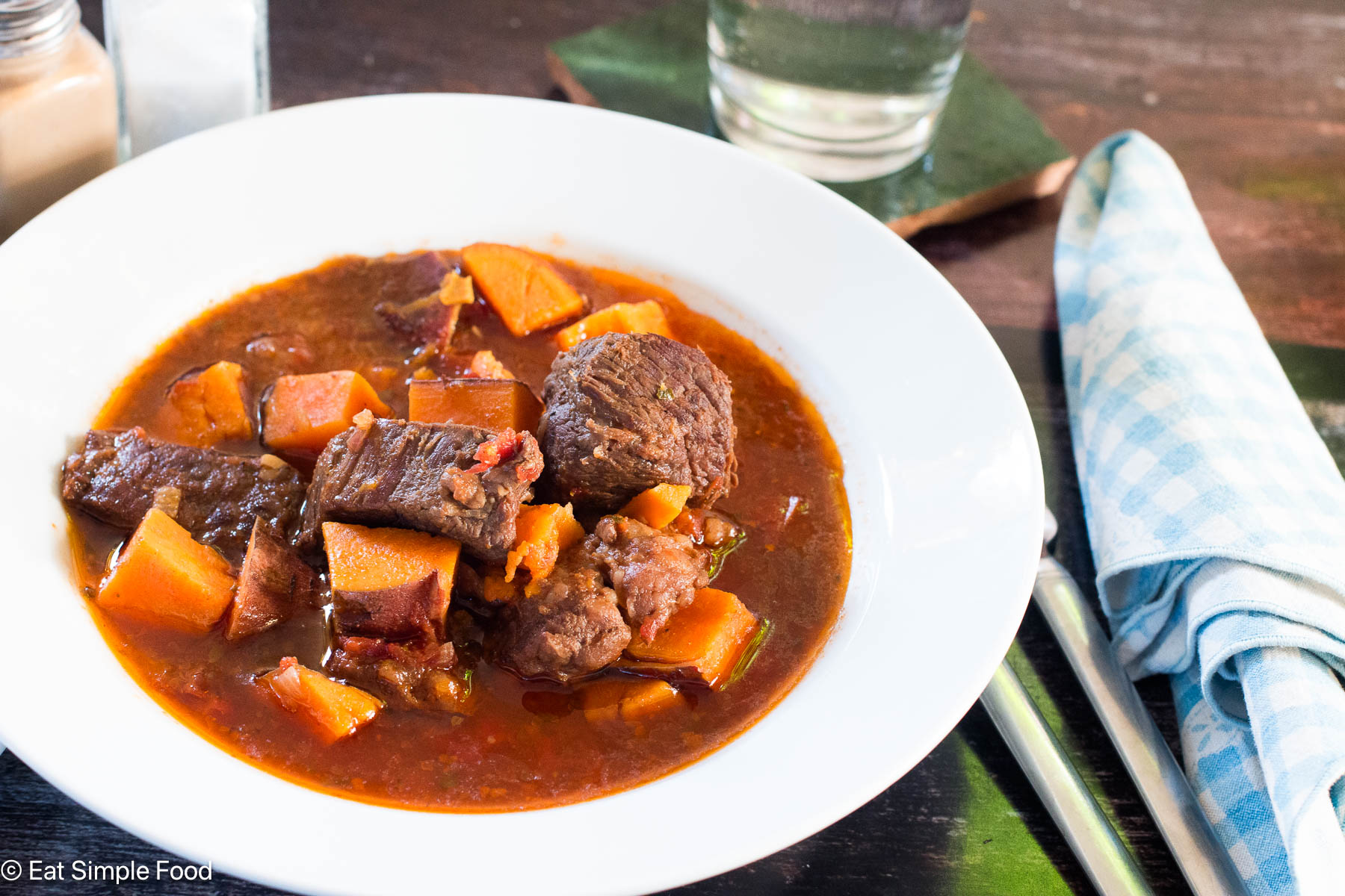 Side view of white bowl of brown stew with chunks of orange sweet potatoes. Napkin, glass of water, and silverware on the side.