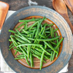 Brown Tan plate of cooked green beans in sauce with wooden utensils on the side.