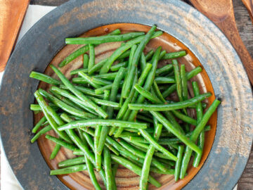 Brown Tan plate of cooked green beans in sauce with wooden utensils on the side.