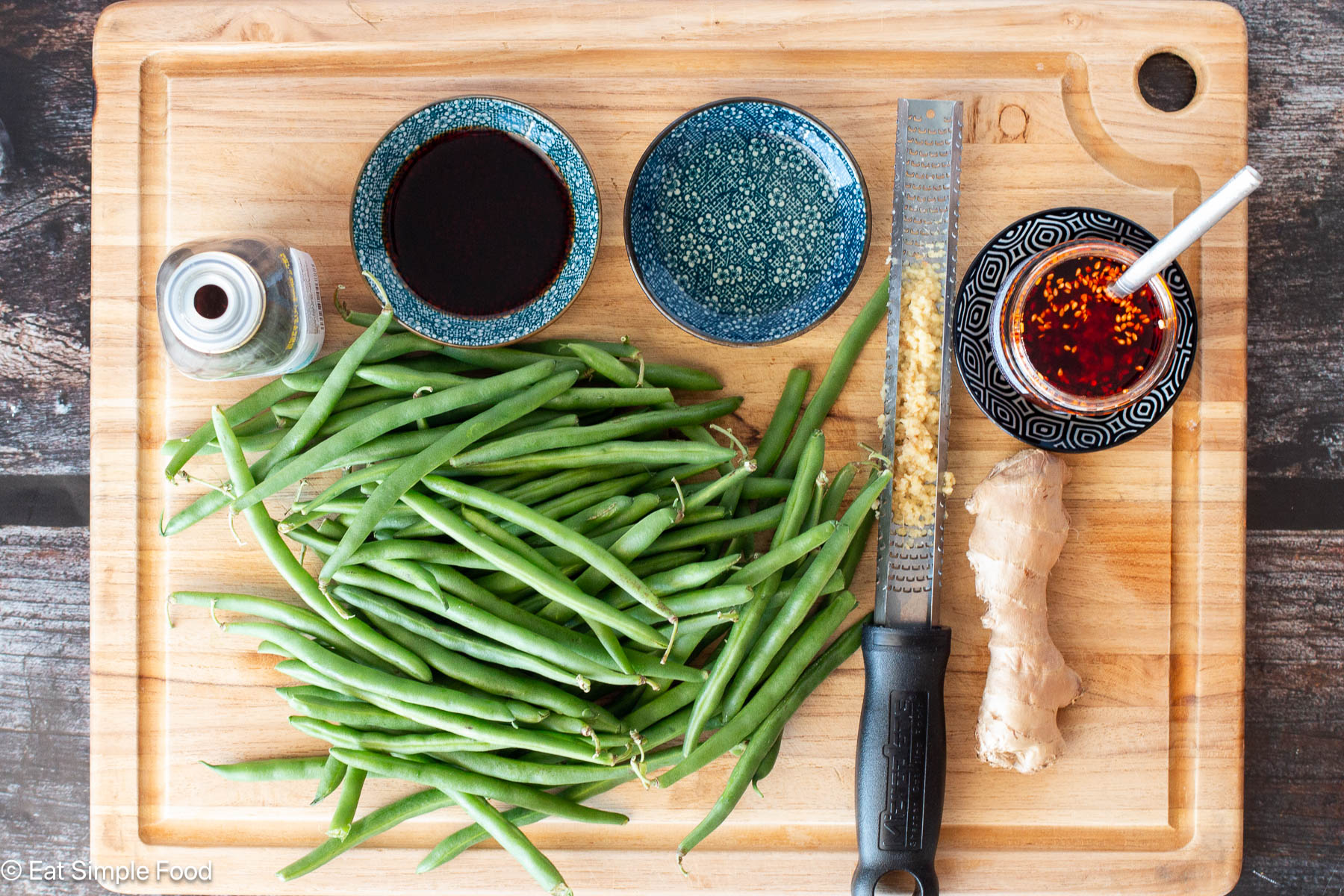 Top down view of wood cutting board with a pile of green beans and a piece of ginger and small containers with sauces.
