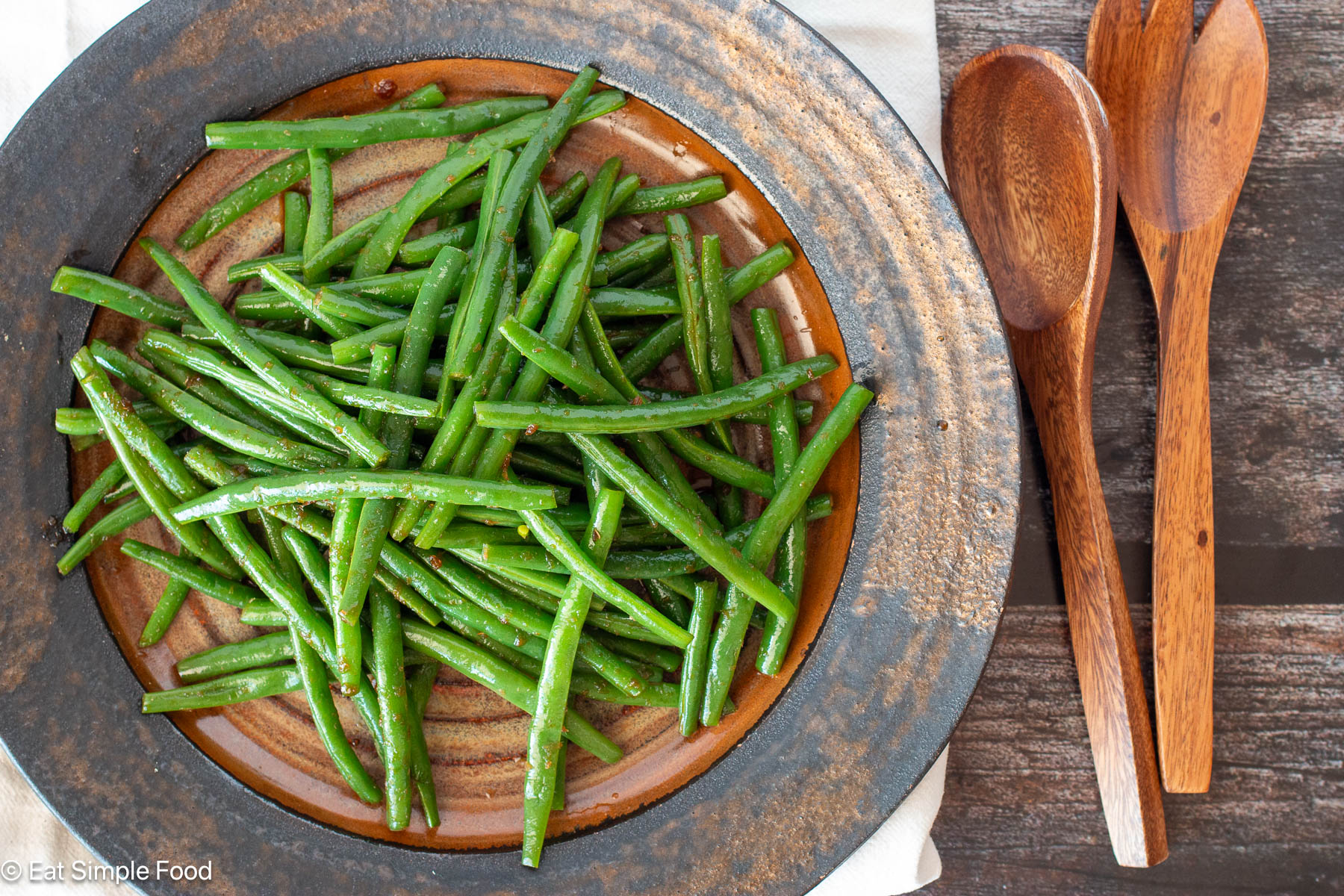 Brown Tan plate of cooked green beans in sauce with wooden utensils on the side on a brown table.