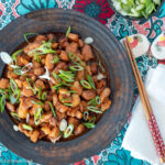 Top view of brown plate with diced chicken in brown sauce and sliced green onions on top. chopsticks on the side.