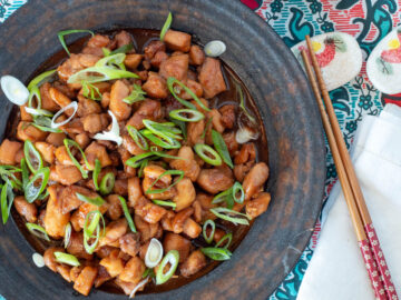 Top view of brown plate with diced chicken in brown sauce and sliced green onions on top. chopsticks on the side.
