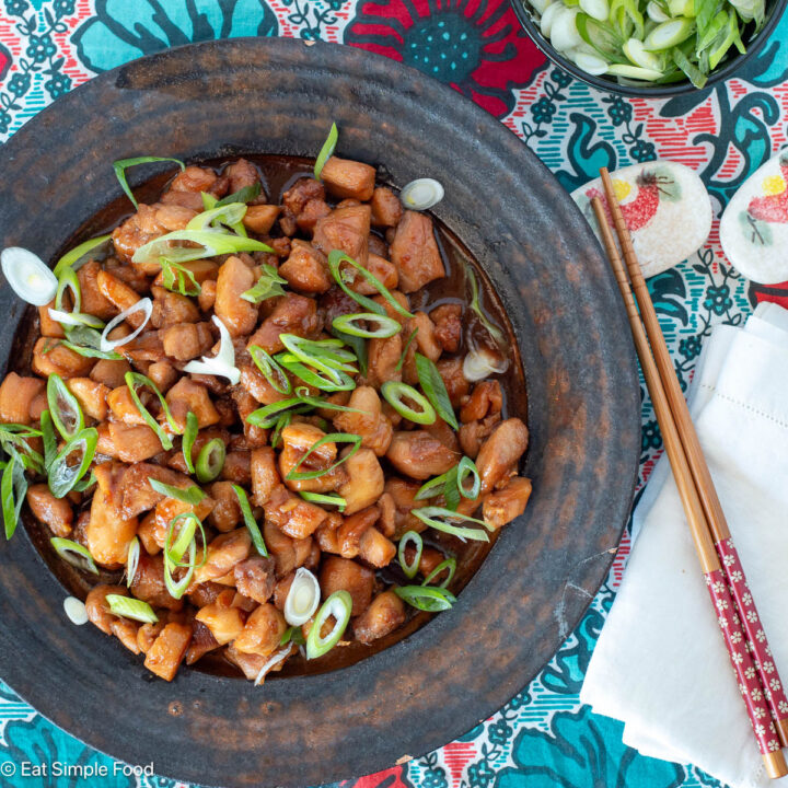 Top view of brown plate with diced chicken in brown sauce and sliced green onions on top. chopsticks on the side.