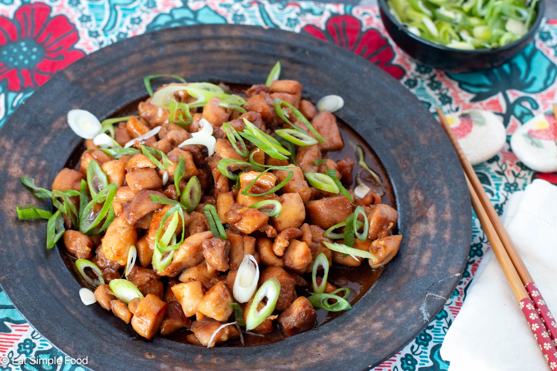Side view of brown plate with diced chicken in brown sauce and sliced green onions on top. chopsticks on the side.