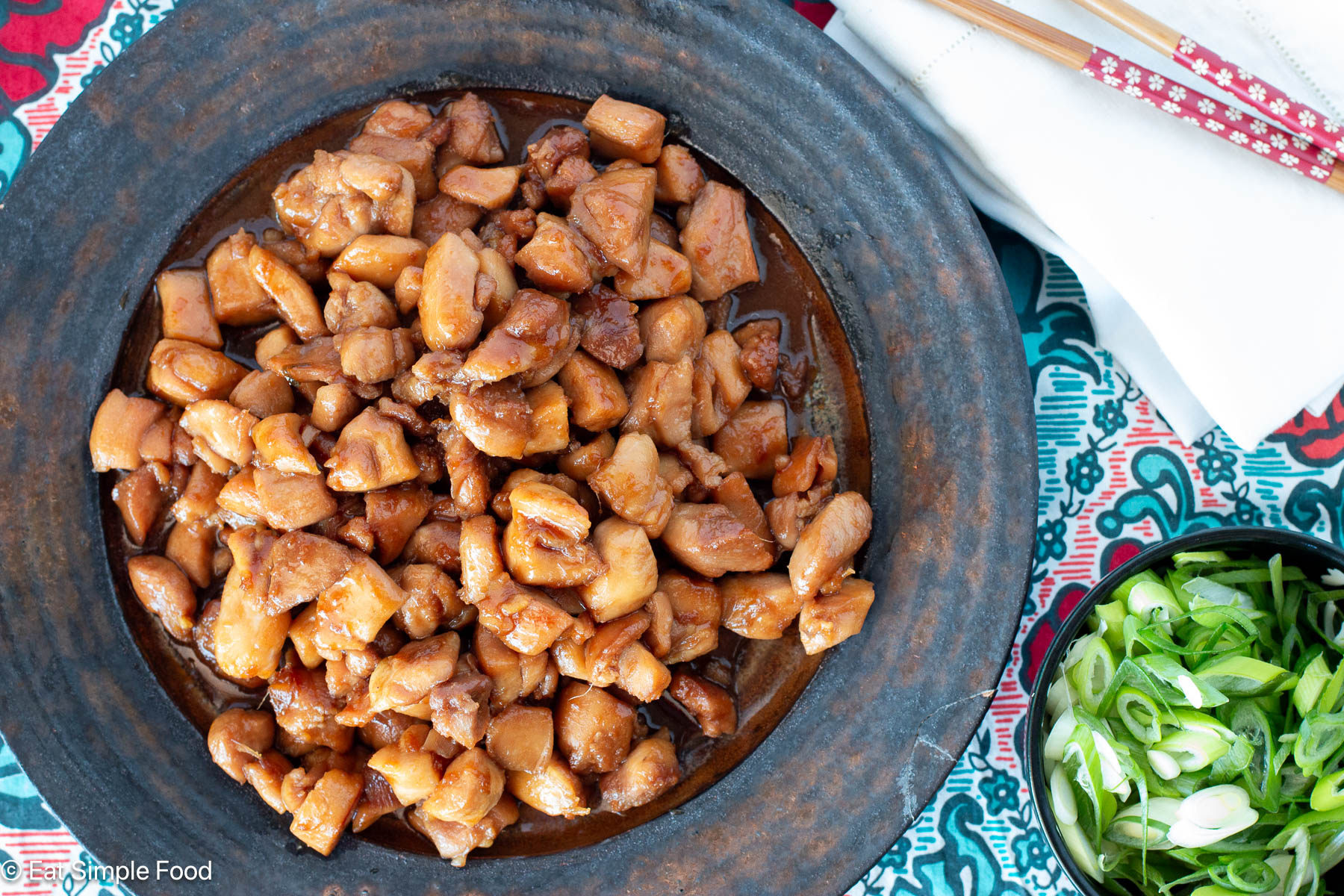 Top view of brown plate with diced chicken in brown sauce and sliced green onions on top. Sliced green onions and chopsticks on the side.