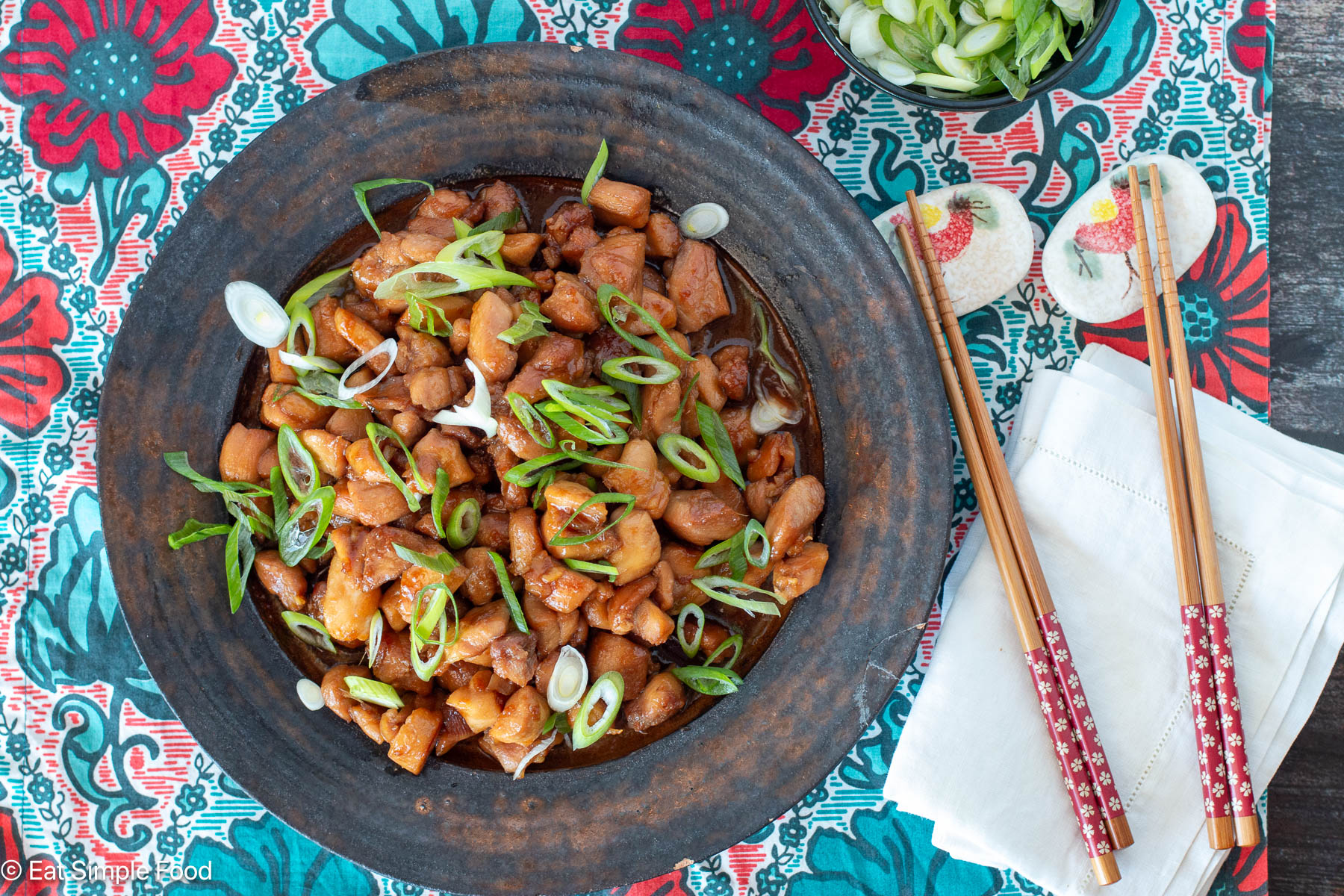 Top view of brown plate with diced chicken in brown sauce and sliced green onions on top. chopsticks, napkins, and green onions on the side.