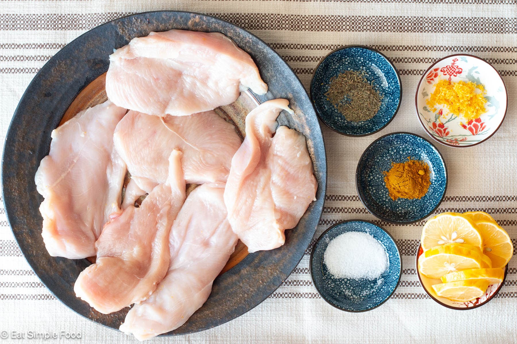 Top down view of a plate of six raw chicken breasts on a brown plate with 5 small ramekins of spices on the sides.