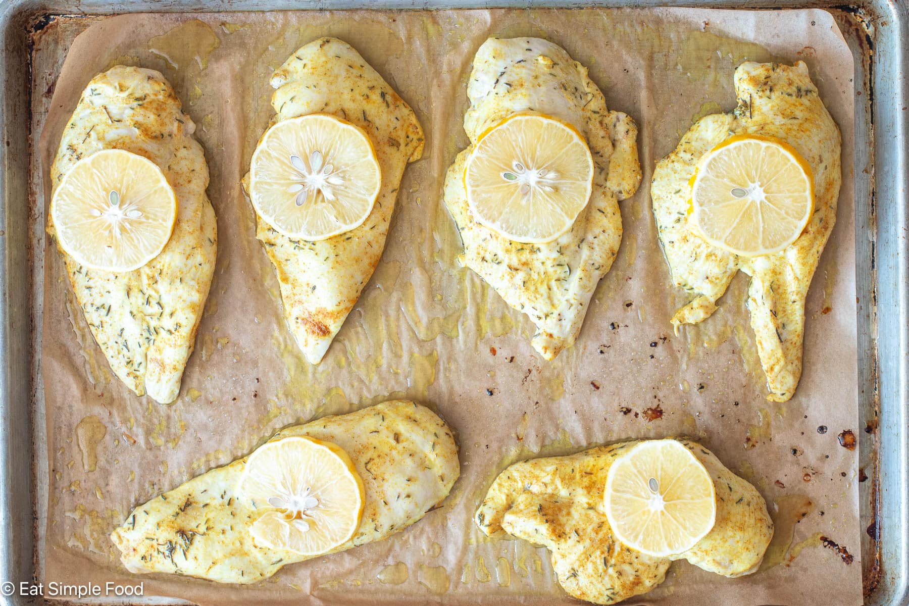 Top down view of six cooked pieces of chicken breast with lemon slices on top on a sheet pan lined with parchment paper.