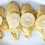 Top down view of 4 pieces of chicken breast with seasoning on top and lemon slices on a white plate. Close up.