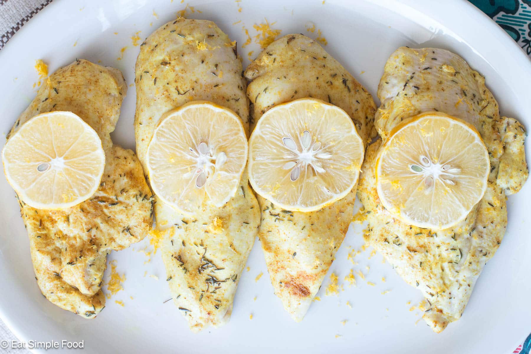Top down view of 4 pieces of chicken breast with seasoning on top and lemon slices on a white plate.