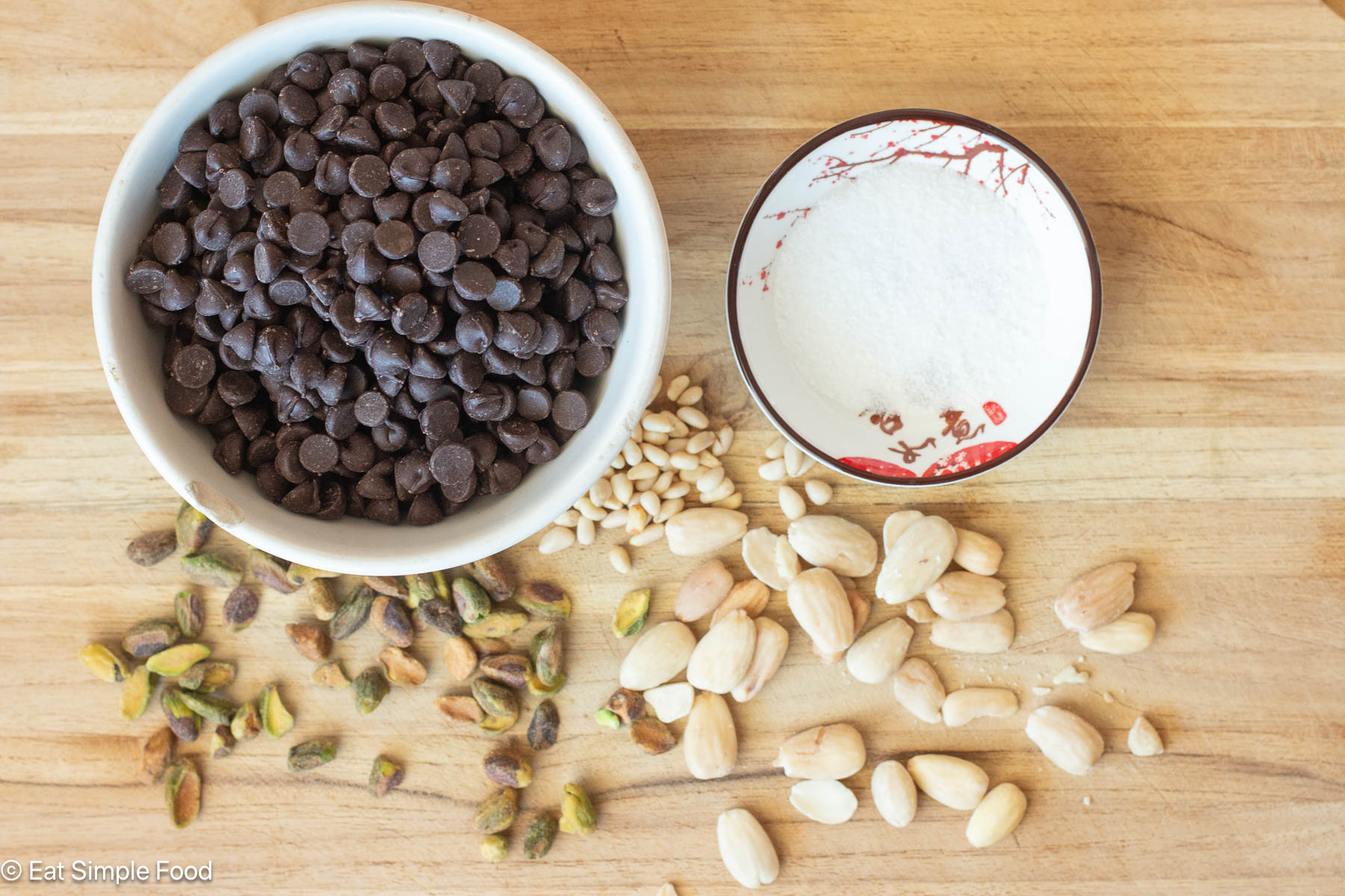Top down view of bowl of chocolate chips, sea salt, and pistachios, almonds, and pine nuts on a wood cutting board.