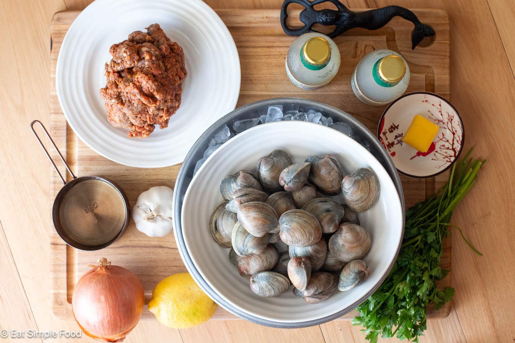Top down view of clams on ice with a whole lemon, whole onion, whole garlic bulb, tab of butter, bunch of parsley and white plate of raw sausage.