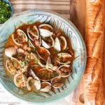 Top down view of open clams and sausage in sauce in a green bowl with a baguette on the side.