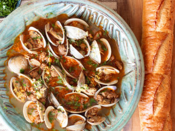 Top down view of open clams and sausage in sauce in a green bowl with a baguette on the side.