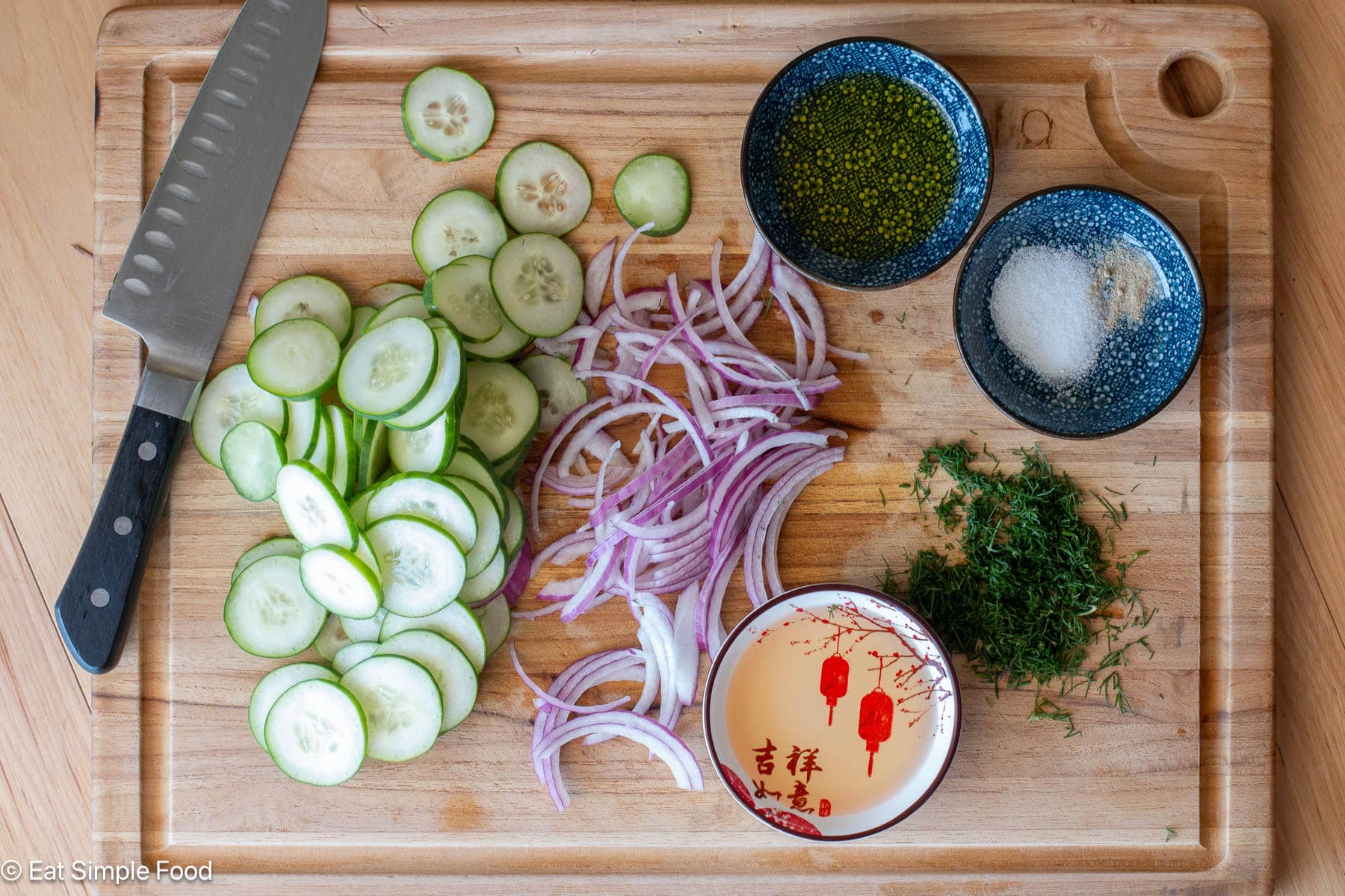 Ingredients on a wood cutting board: sliced cucumbers, sliced red onions, chopped herbs and small containers of liquid ingredients. Top down view.