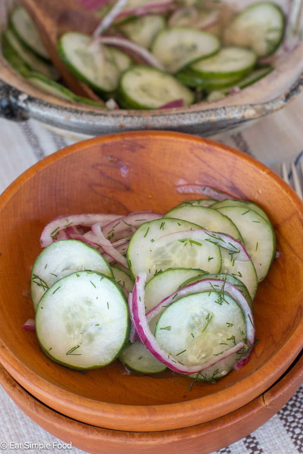 Side view of sliced cucumbers and red onions in a wood bowl with a larger bowl of the same behind it.