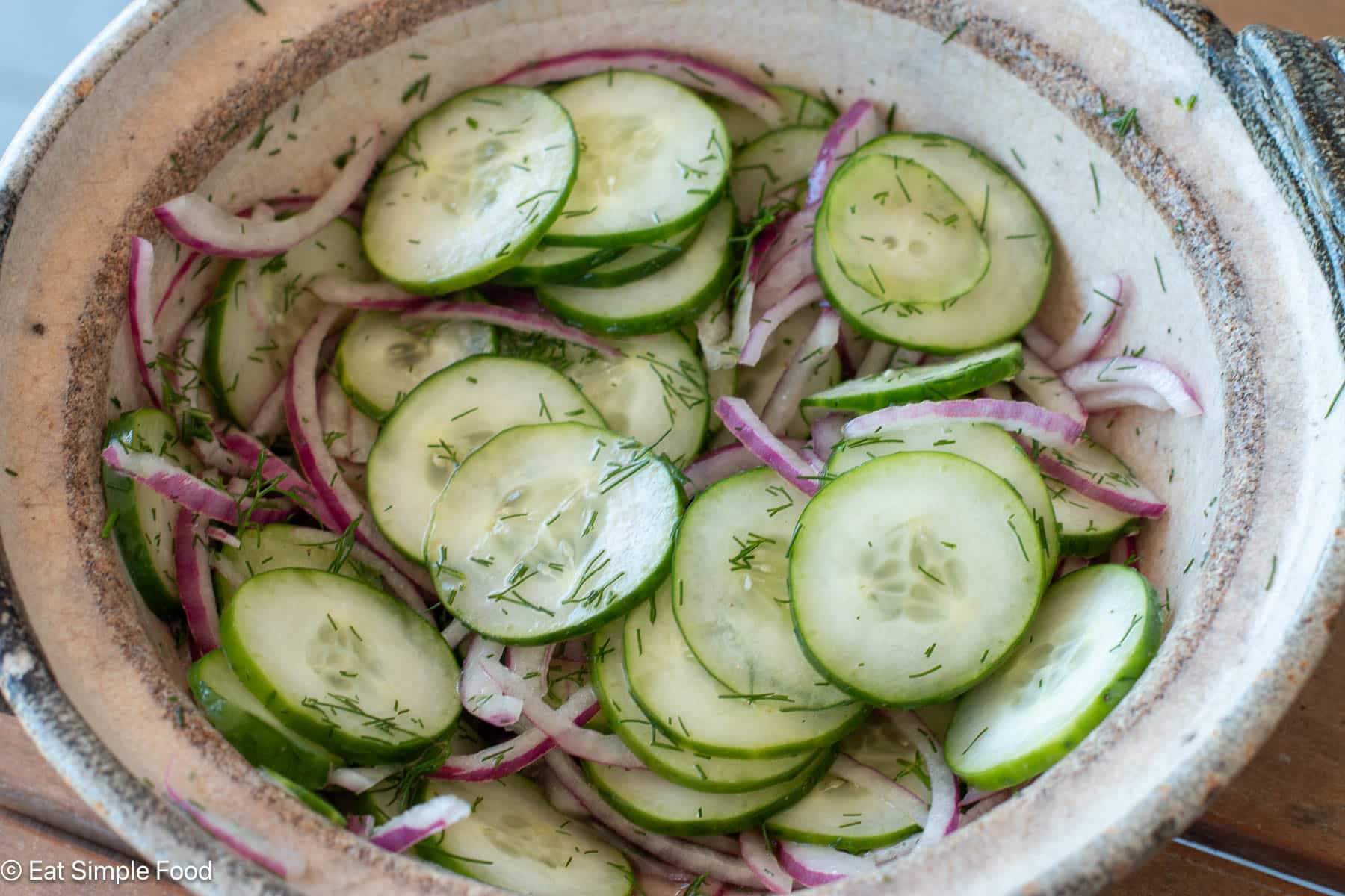 Side view of sliced cucumbers and red onions in a brown bowl.
