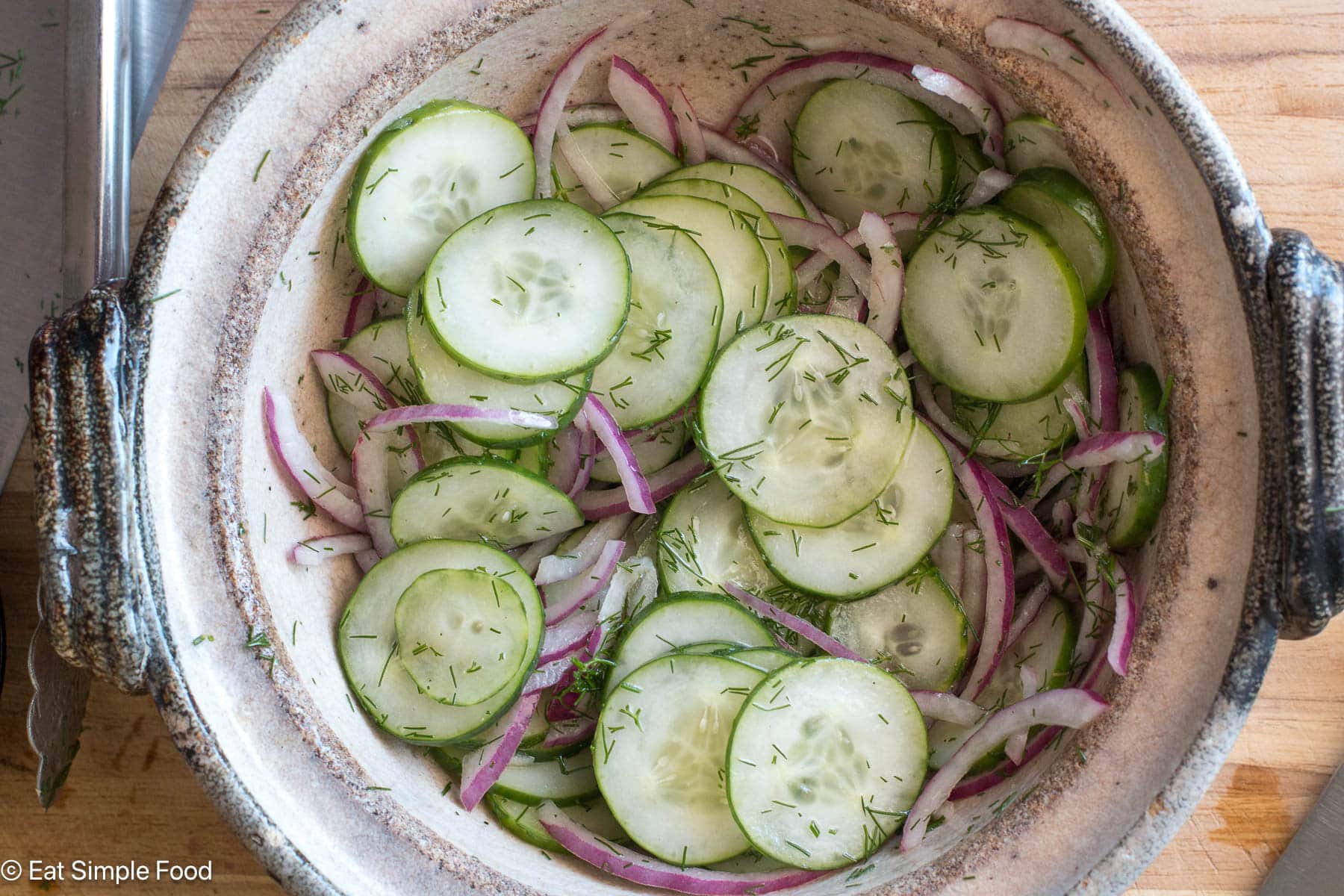 Top down view of sliced cucumbers and red onions in a brown bowl.