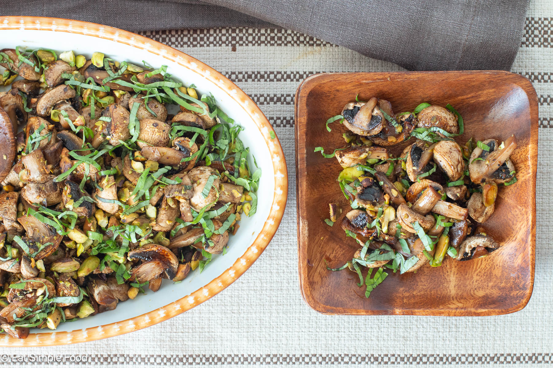 Top down view of browned mushrooms on a brown plate with a large oval white bowl of cooked mushrooms on the side. Brown napkins in background.