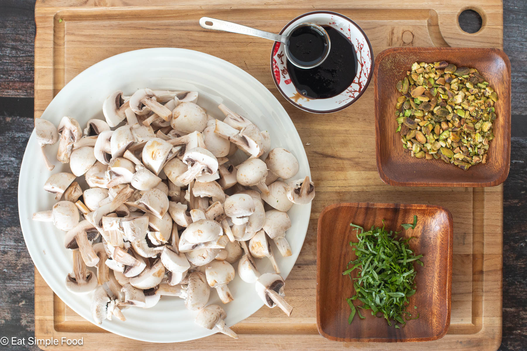 Top down view of quartered raw mushrooms on a white plate thats on a wood cutting board. Pistachios and brown sauce, and chopped green garnish on the side.