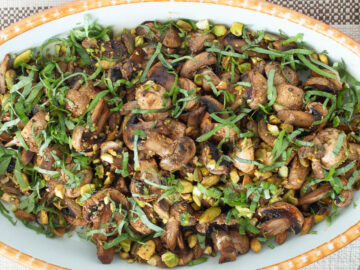 Top down view of white oval bowl filled with cooked mushrooms with pistachios and chopped basil. Close up.