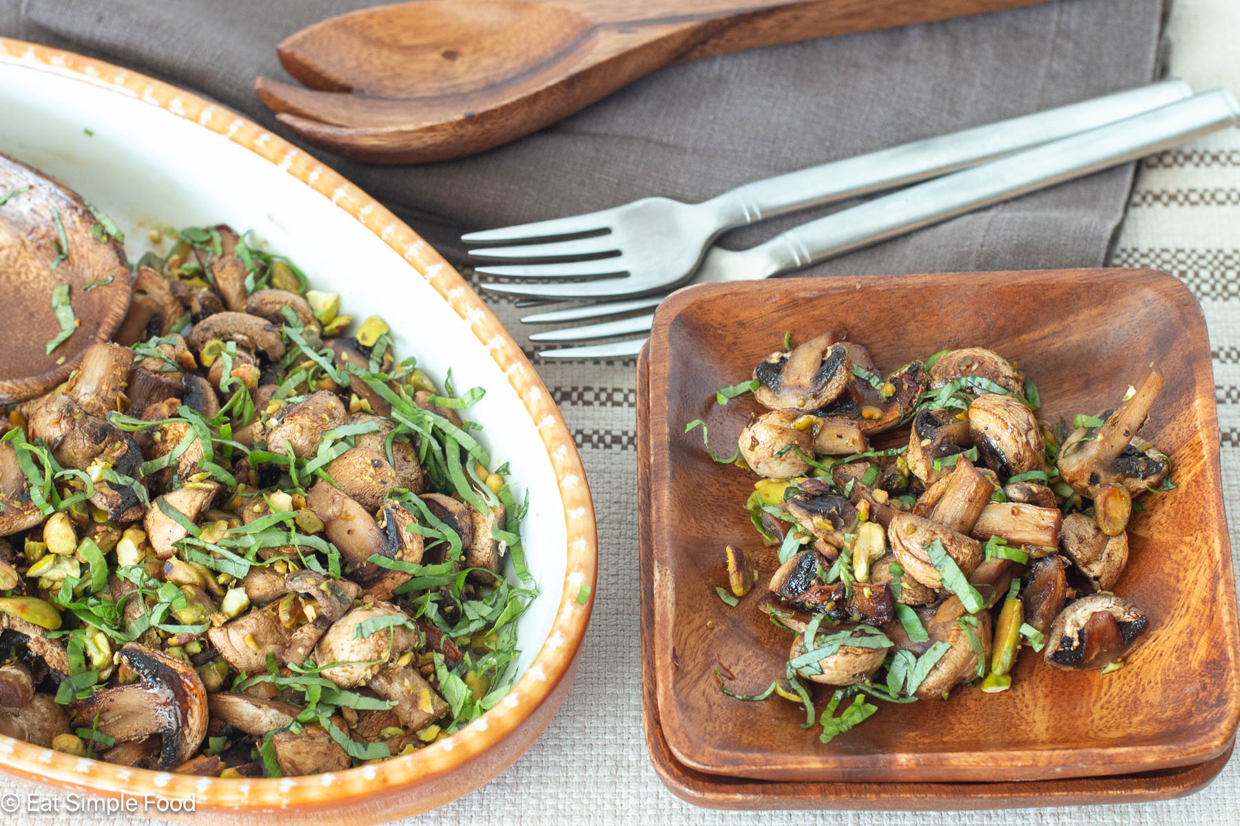 Side view of brown plate with cooked mushrooms and larger oval bowl on the side with cooked mushrooms. Two forks in the background.