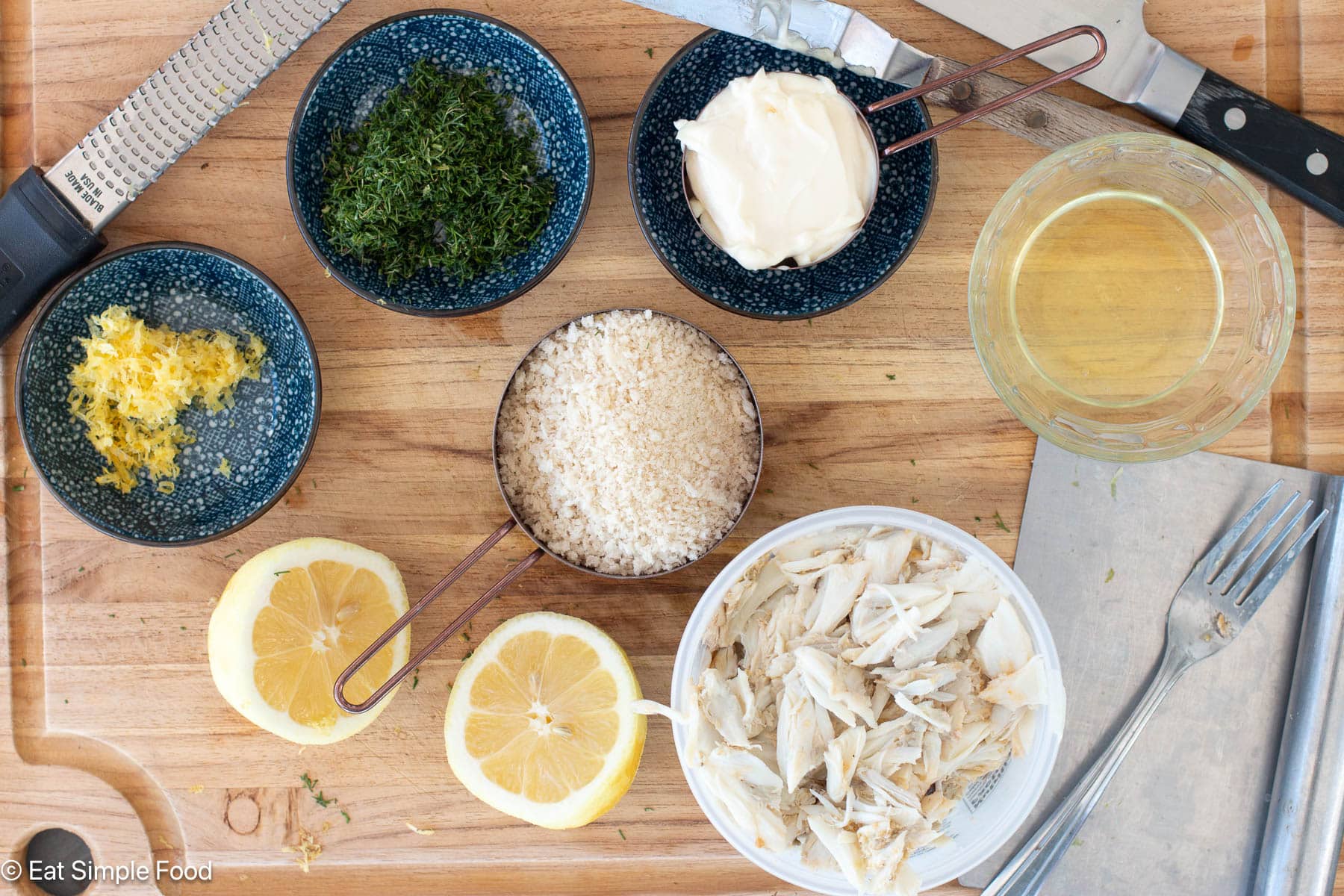Ingredients on a wood cutting board with chefs knife and fork: crab, bread crumbs, lemon halves, chopped dill, lemon zest and mayonnaise.