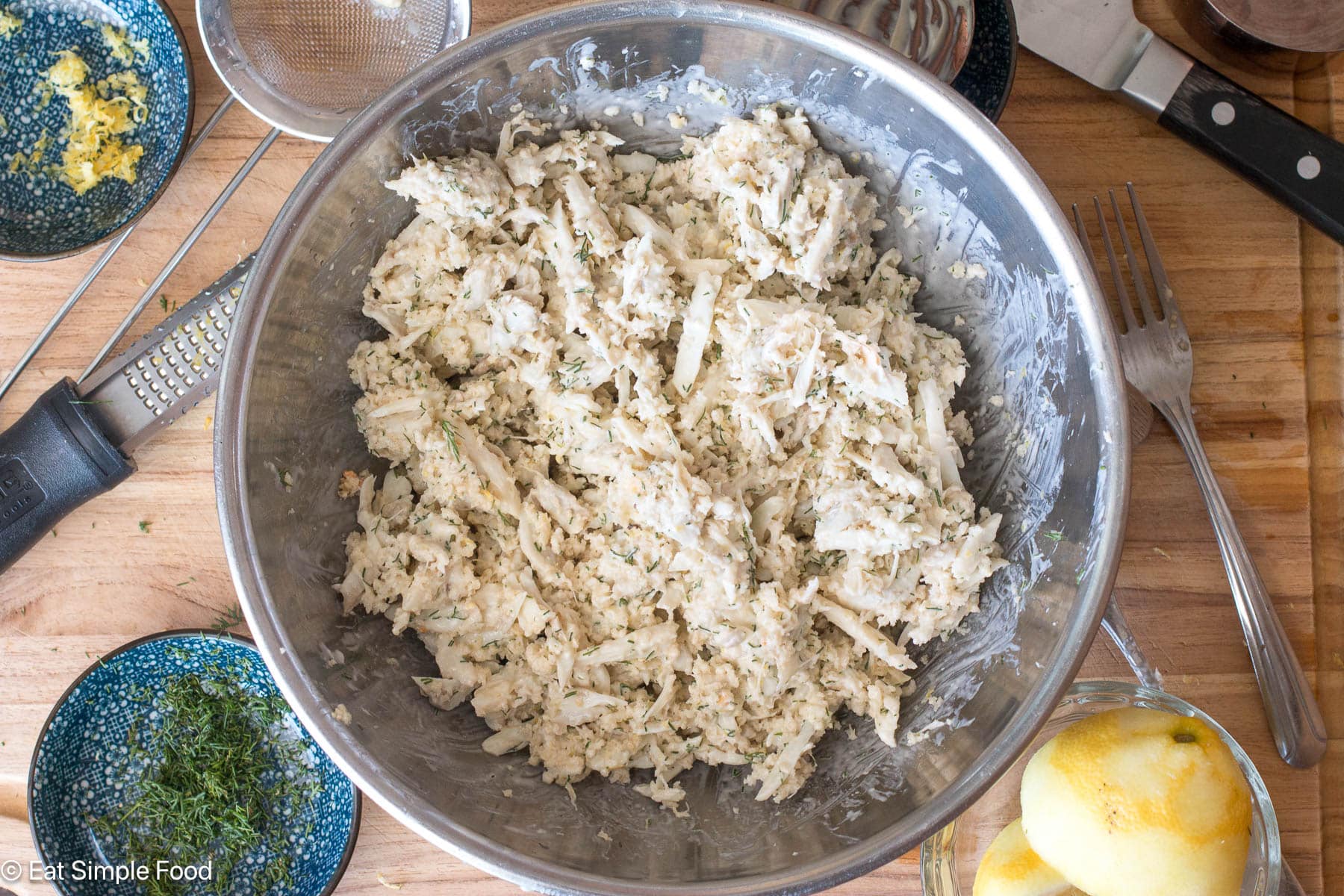 Stainless steel bowl of raw crab mixture on a wood cutting board with lemon and dill on the side with a fork and chefs knife.