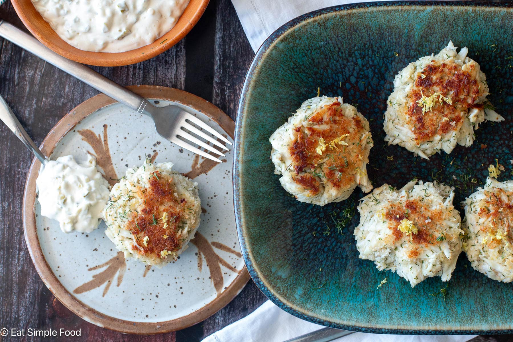 top down view of a plate with a crab cake and white sauce with aqua plate with three crab cakes.