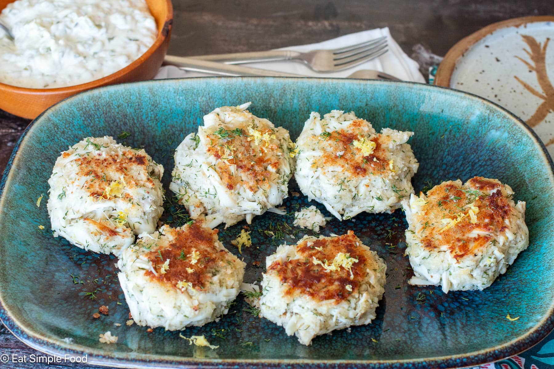 Side view of seared crab cakes on an aqua rectangle plate. Bowl of white sauce in background.