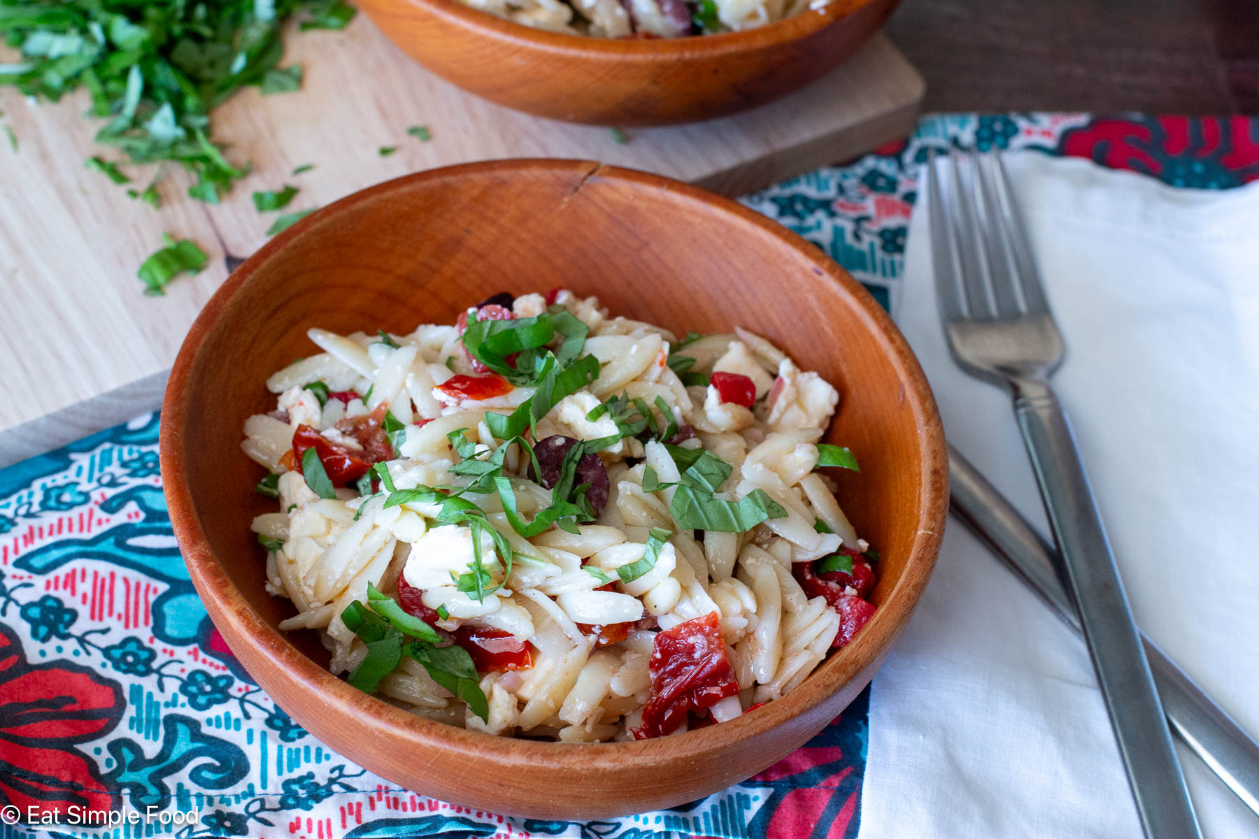 Side view of a wood bowl of pasta salad with basil and silverware on the side.