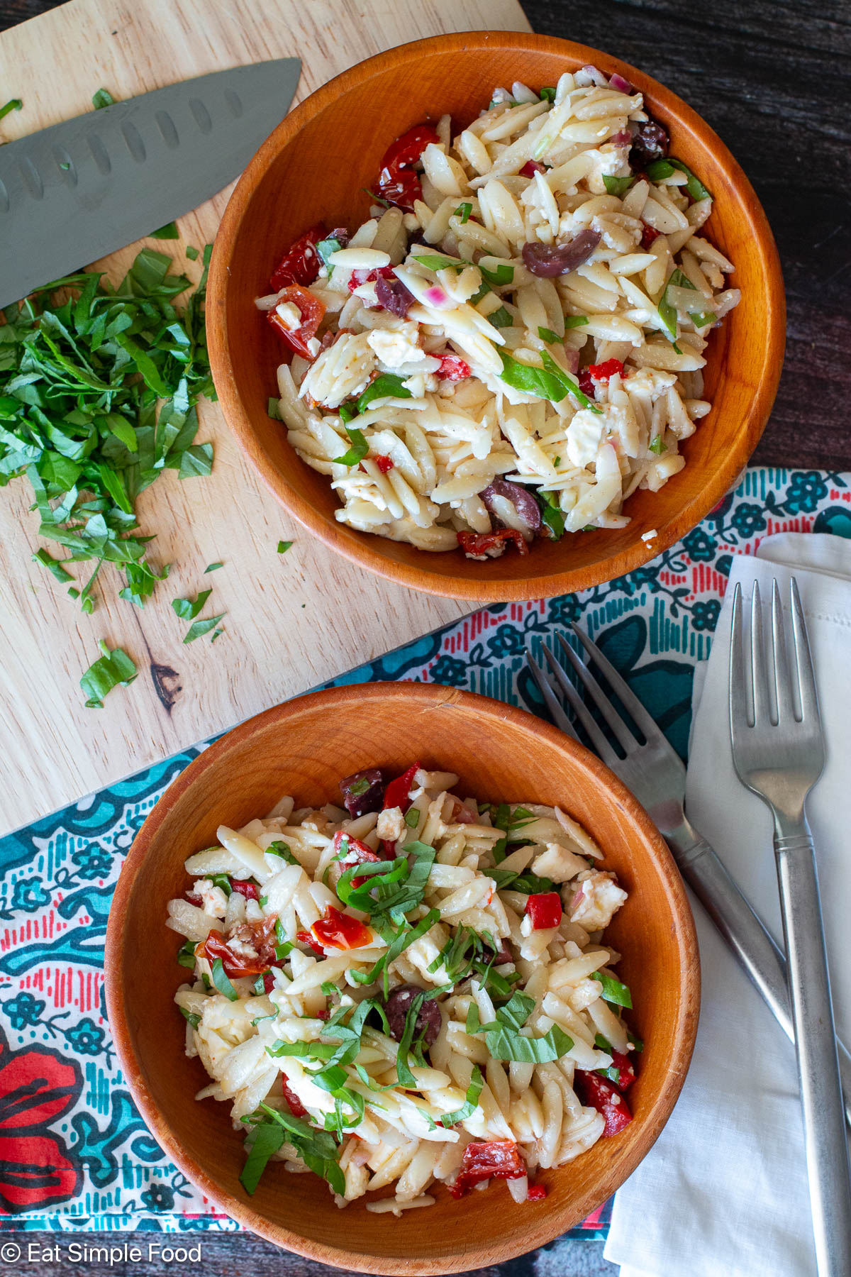Top down view of two wood bowl filled with pasta on a colorful tablecloth. chefs knife in background with basil.